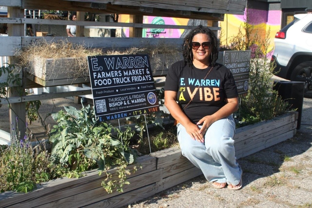 A woman sits outside a business on a the edge of a planter, wearing an "E. Warren: It's a vibe" t-shirt and posing next to a sign for the E. Warren Farmers Market