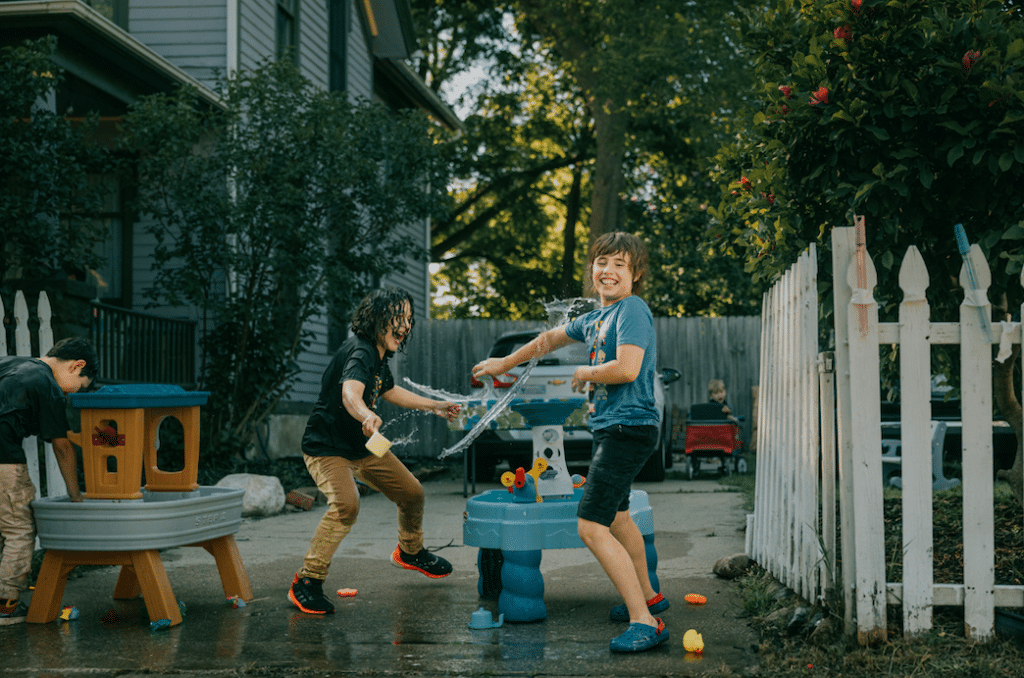 Several children playing with water tables in a driveway, splashing each other and smiling.