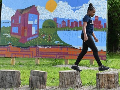 A young African American girl walks across a row of tree stumps in front of a mural. The mural features a city and a house, as well as depictions of the water cycle.