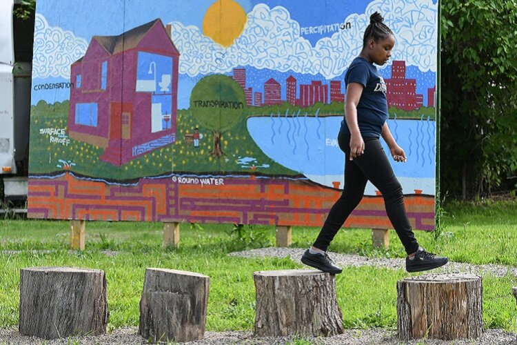 A young African American girl walks across a row of tree stumps in front of a mural. The mural features a city and a house, as well as depictions of the water cycle.